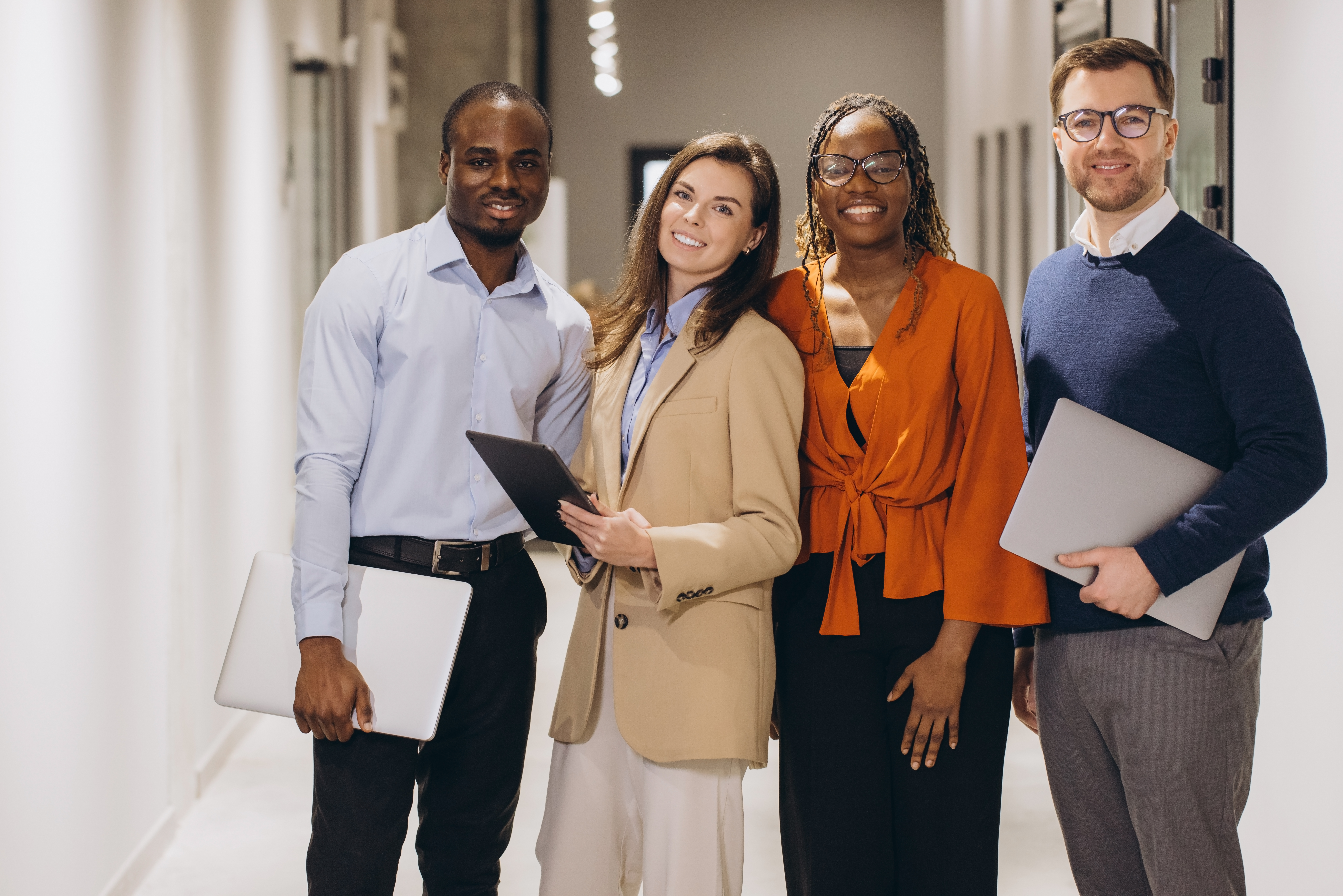 Multi Ethnic Businesspeople Standing Together In Office Hallway Holding Laptops And Digital Tablet, Smiling